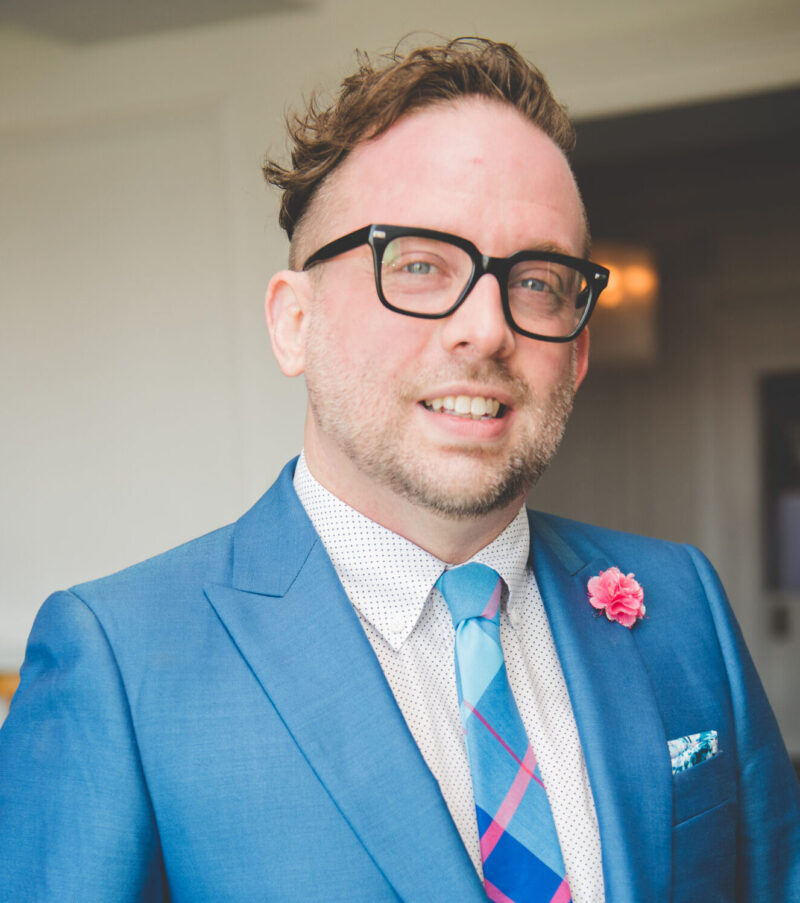 A man in blue suit and glasses smiling for the camera.